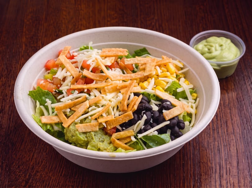 A vibrant salad bowl with lettuce, corn, black beans, guacamole, cheese, and tortilla strips.