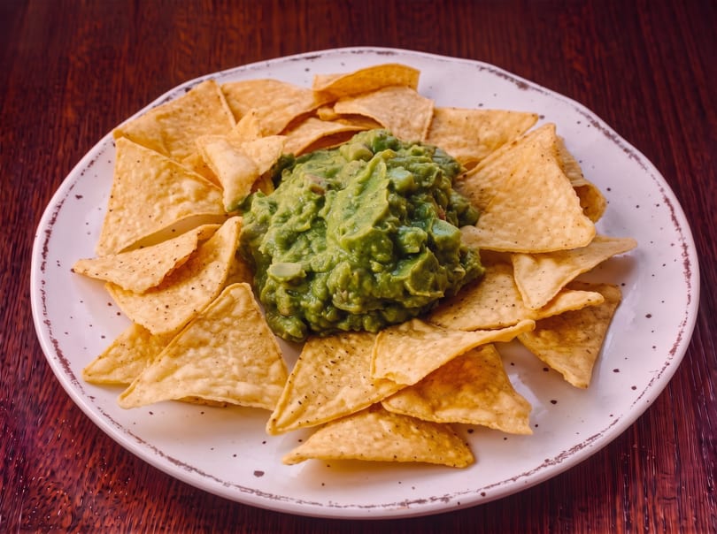 A white plate filled with golden tortilla chips surrounding a generous serving of green guacamole.