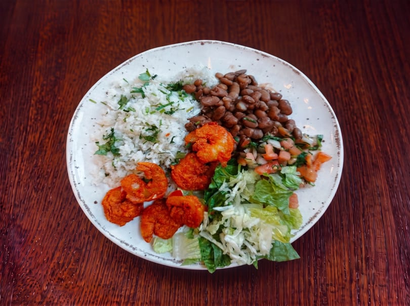 A plate of fried shrimp, rice, beans, pico de gallo, and lettuce on a wooden table.