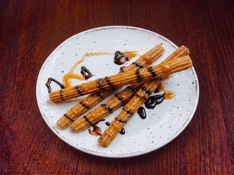 Churros drizzled with chocolate and caramel sauce on a white speckled plate.